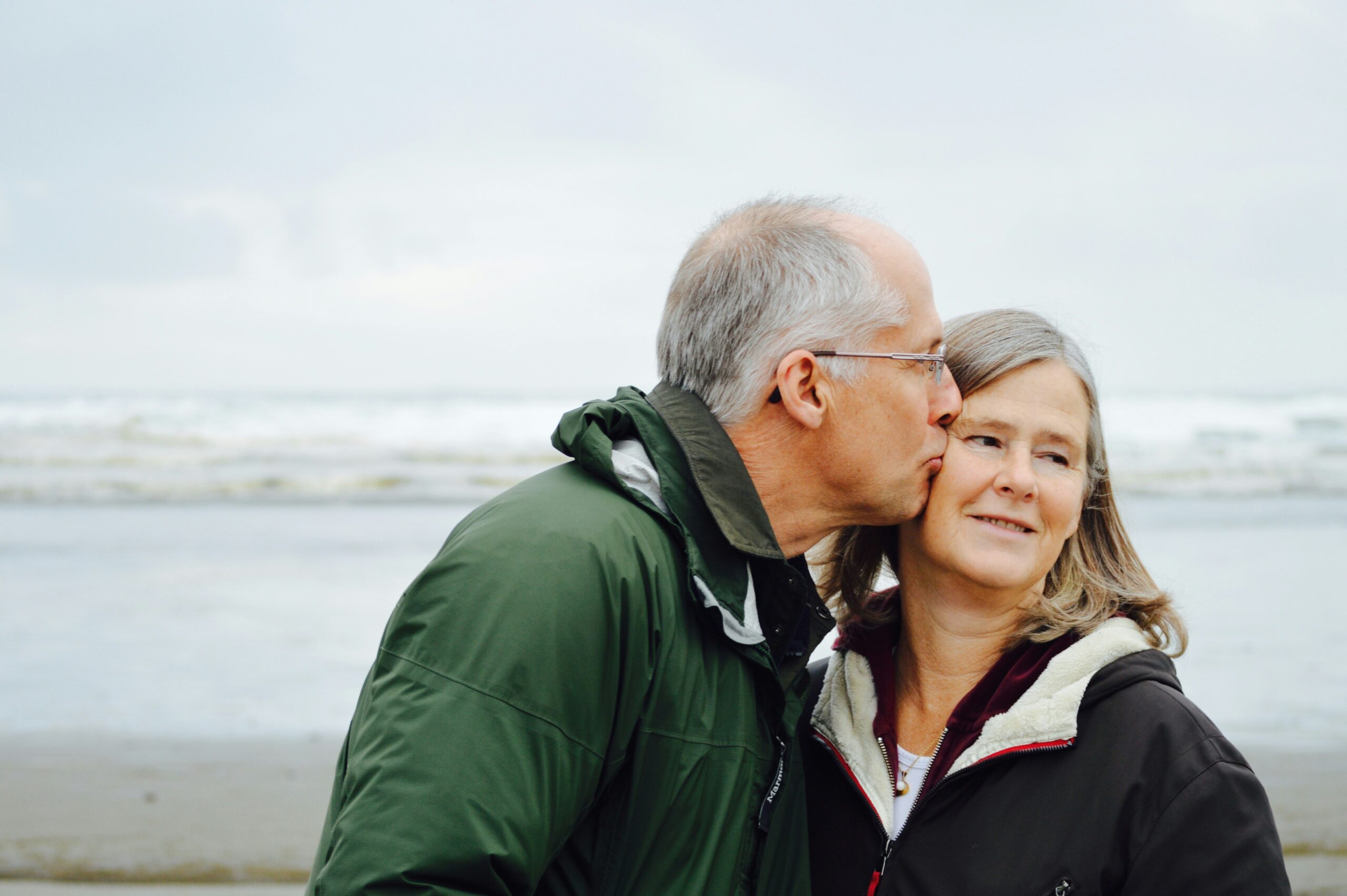 elderly man and woman standing at the beach. Man kissing woman on the cheek.